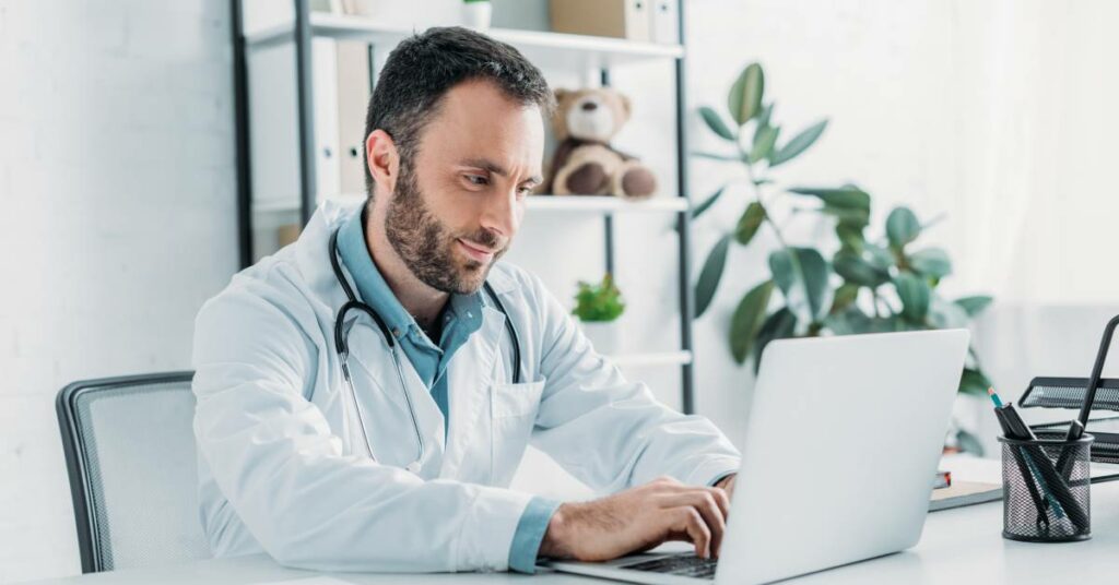 a doctor with a small smile on his face and a stethoscope around his neck typing on a computer to a virtual medical scribe.